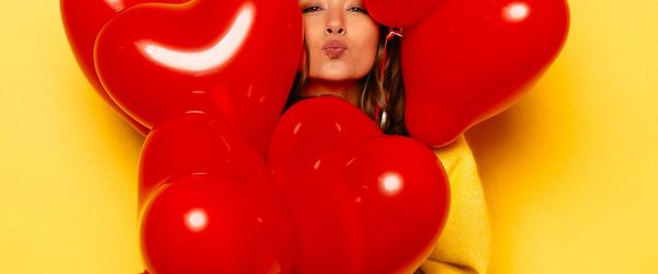 Valentine's day. Smiling girl in yellow sweater giving a kiss, looking out of bunch of red balloons. Isolated over yellow background.